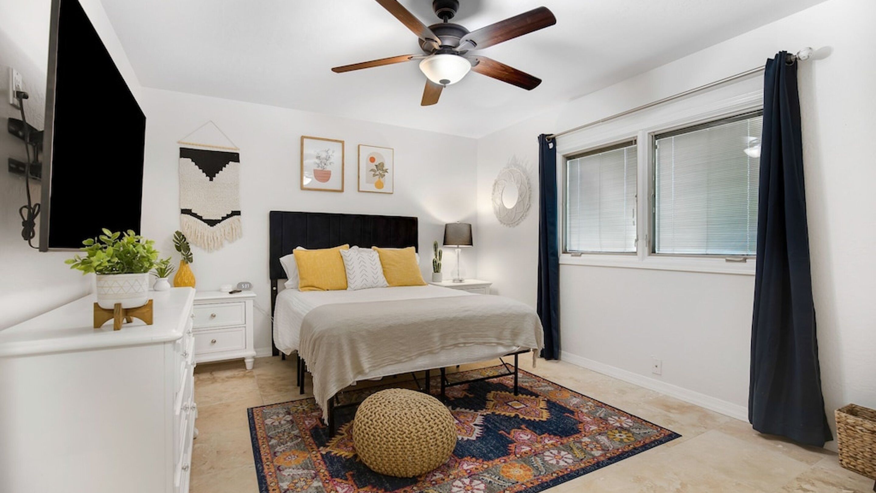 Une chambre à coucher comprenant un lit, des tables de chevet, un ventilateur de plafond, une fenêtre avec des rideaux et un miroir fixé au mur.