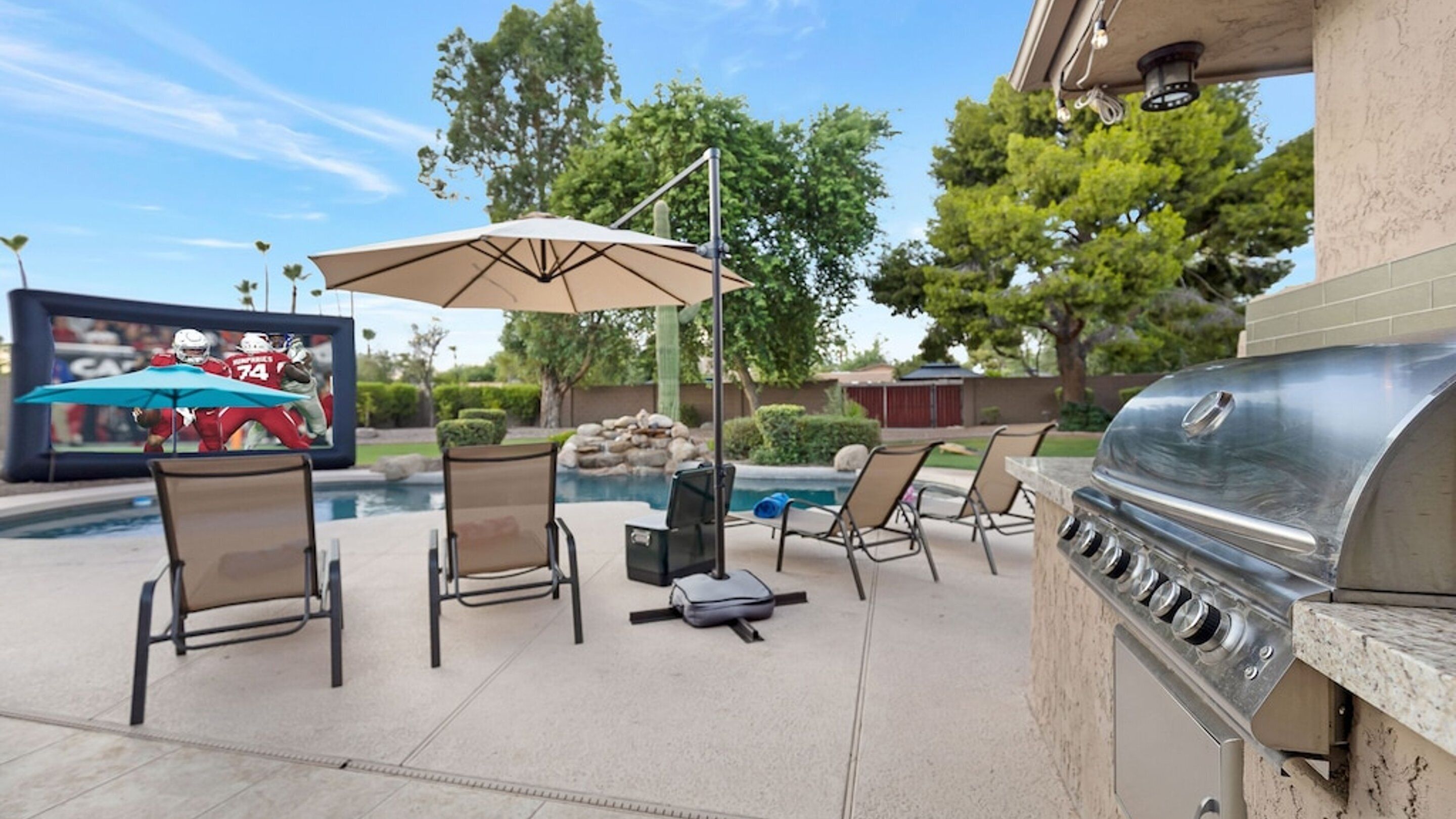 An outdoor patio with a grill, lounge chairs, a pool, and a TV screen displaying a football game.