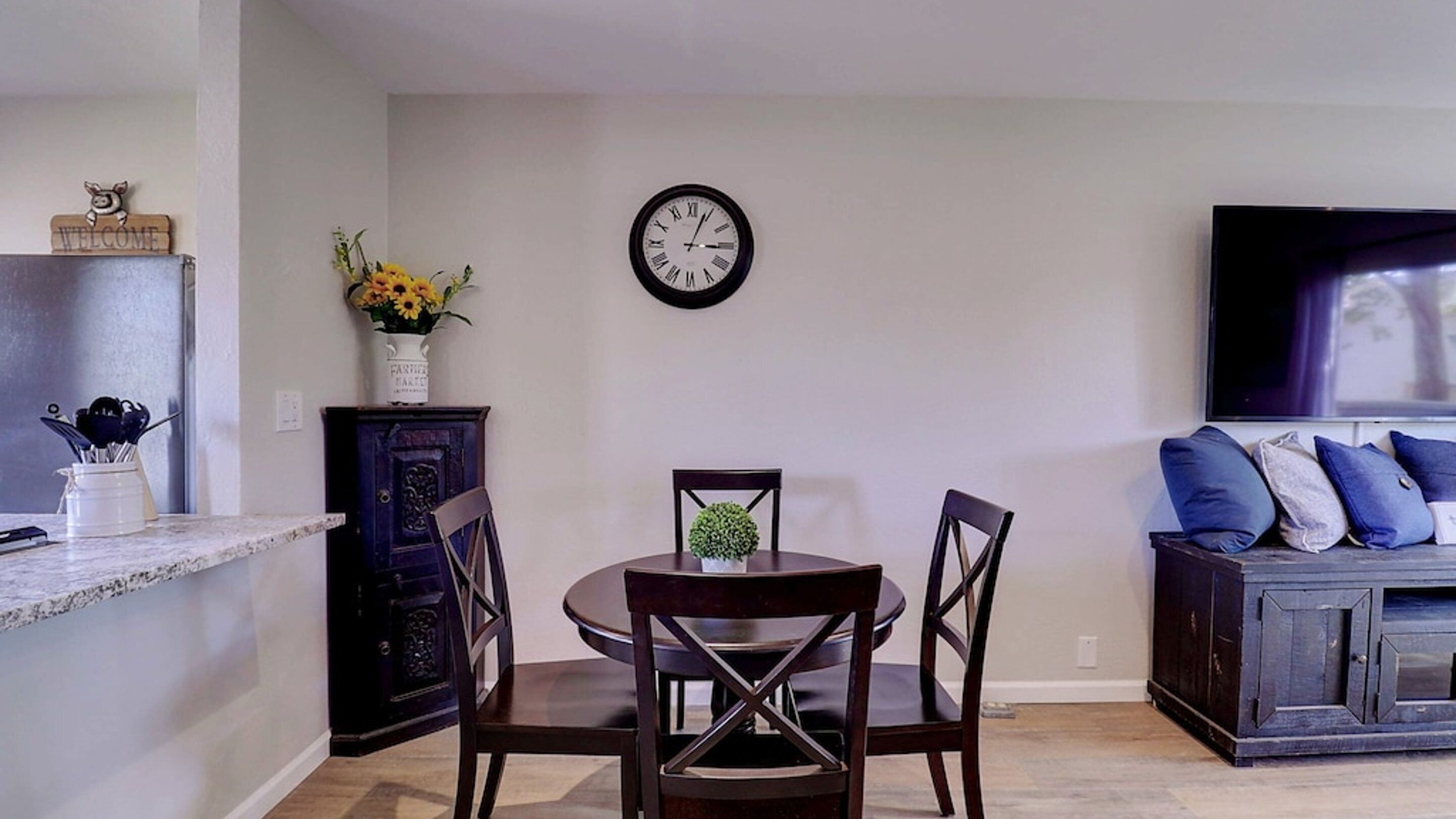 Un coin repas avec une table ronde, deux chaises, une horloge au mur, un vase avec des fleurs et un téléviseur sur le côté droit.