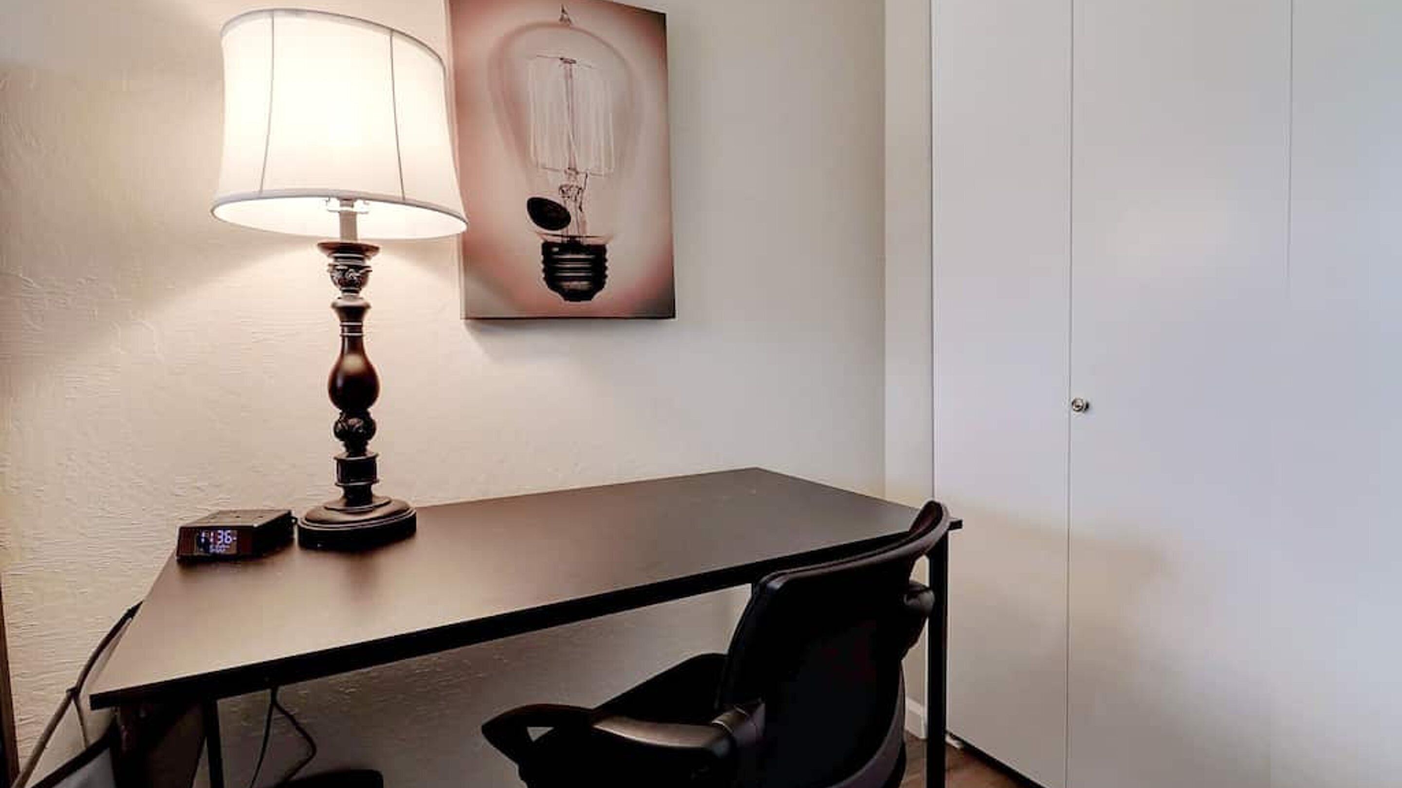 A room with a desk, a chair, a lamp, a clock, and a wall art piece.