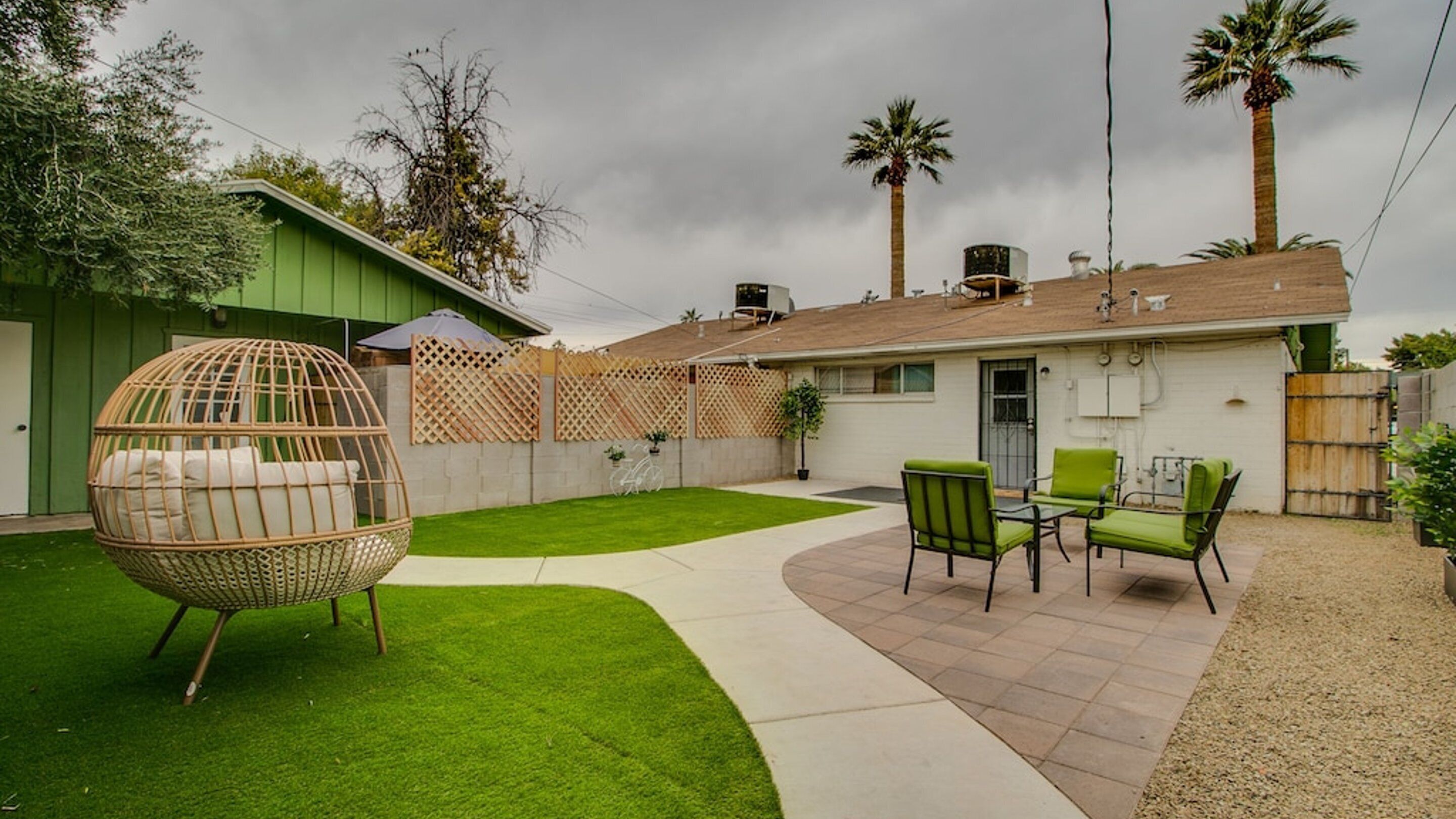 A backyard with a green lawn, a wicker swing chair, and a patio with green chairs.