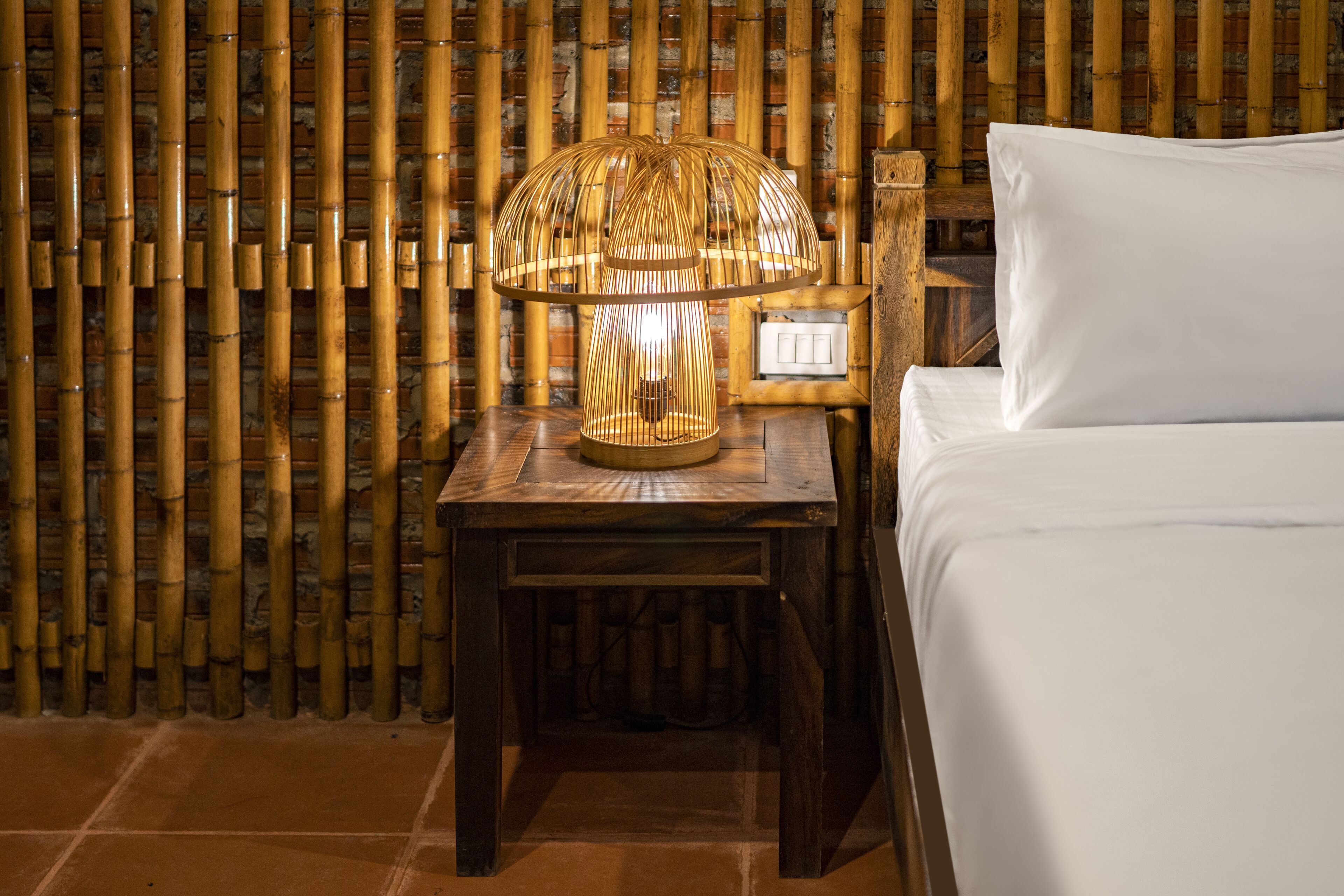 Uma cama com lençóis brancos, uma mesa de cabeceira de madeira com abajur e uma parede de bambu.