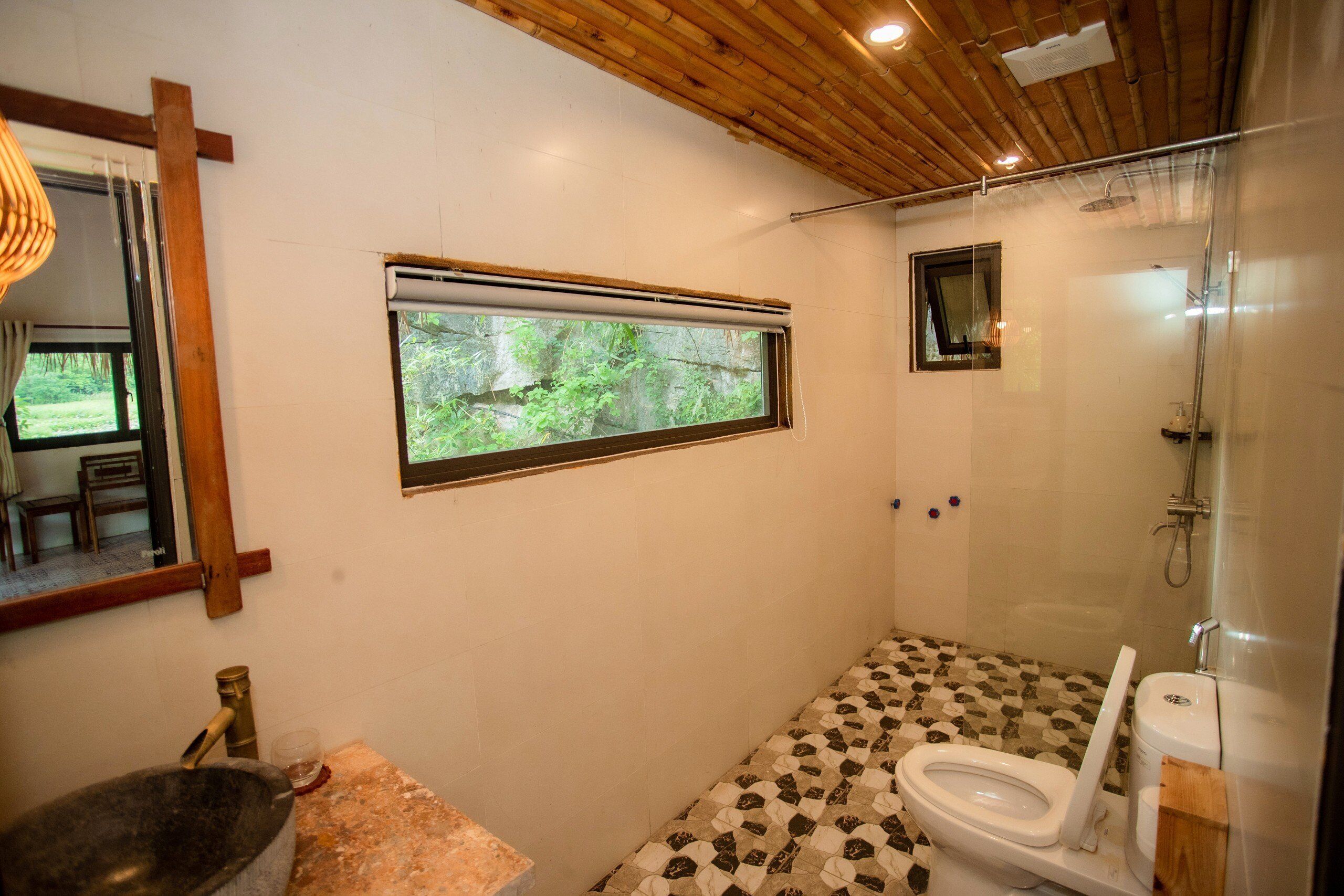 A bathroom with a stone sink, a toilet, a shower, and a window with a view of greenery.