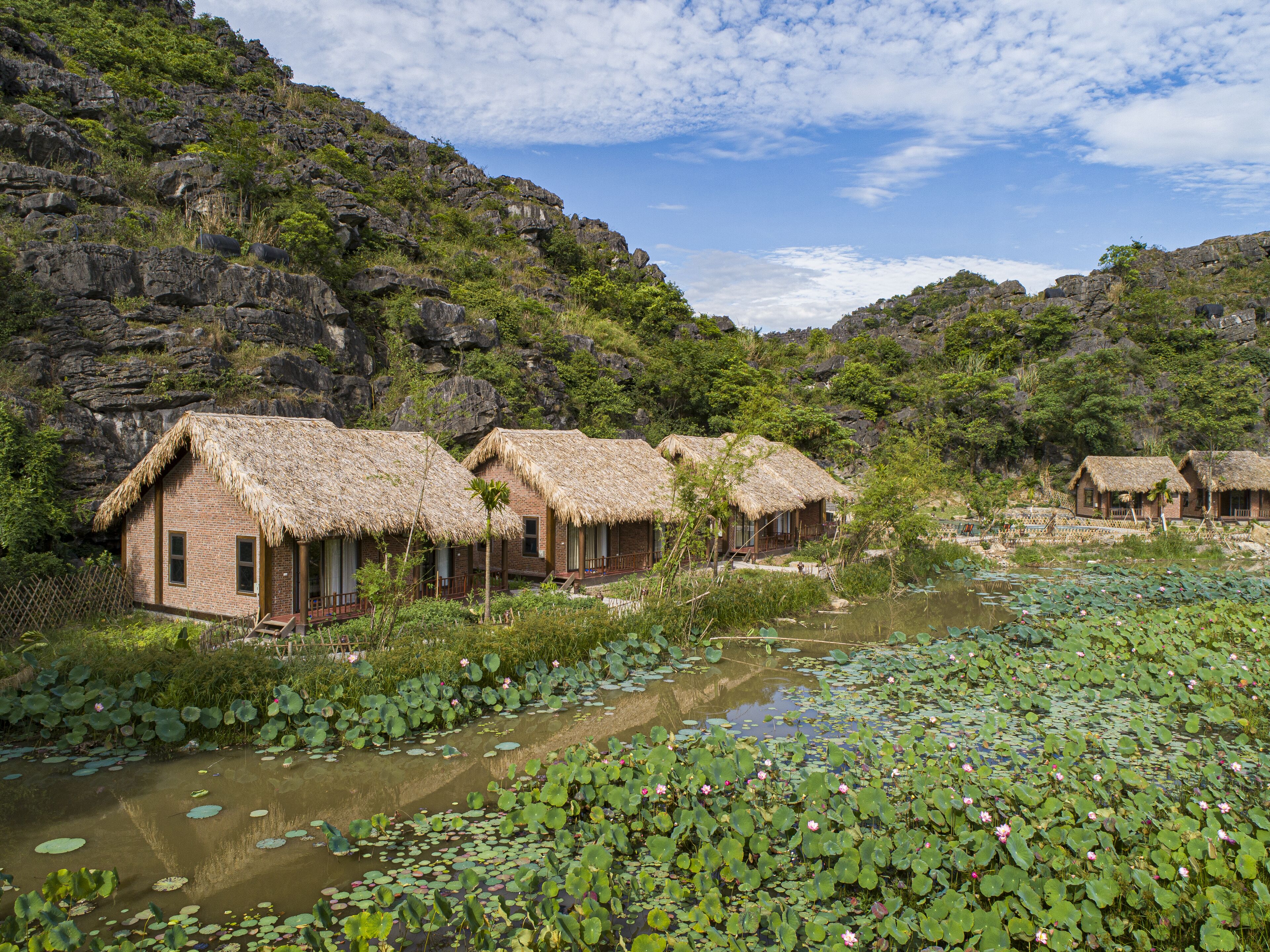 El hotel está ubicado junto a un río con casas de techo de paja, rodeado de exuberante vegetación y colinas rocosas.