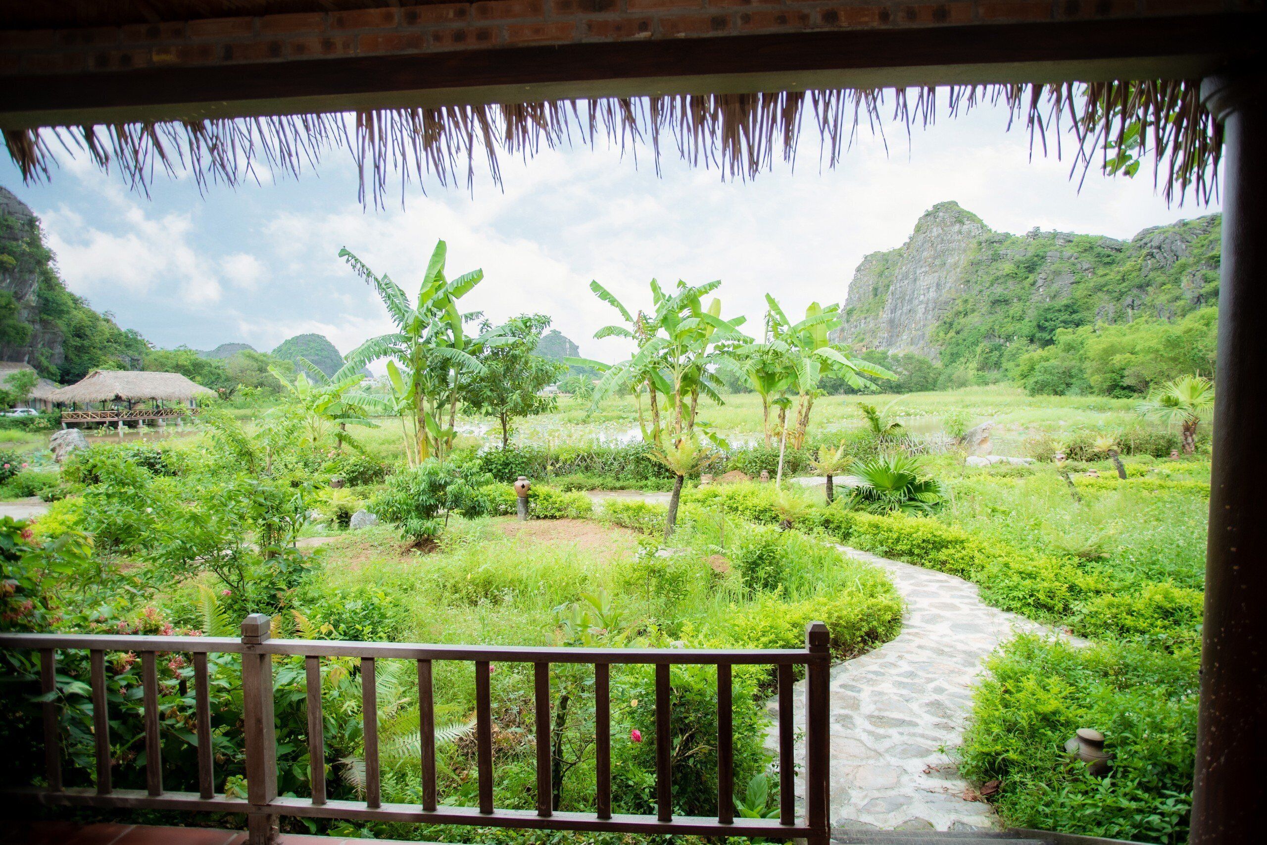 A lush garden with a stone pathway, banana trees, and a thatched-roof structure.