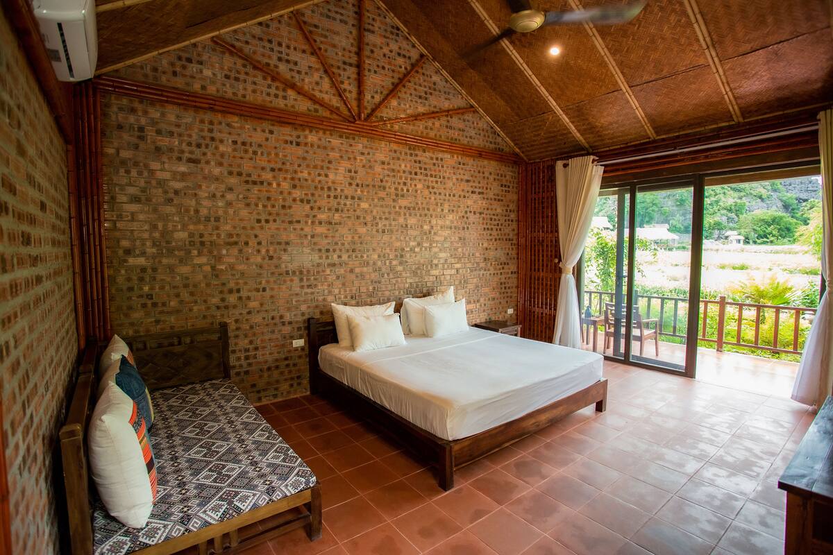 A bedroom with a brick wall, a wooden bed, a wooden bench, and a view of the outdoors.