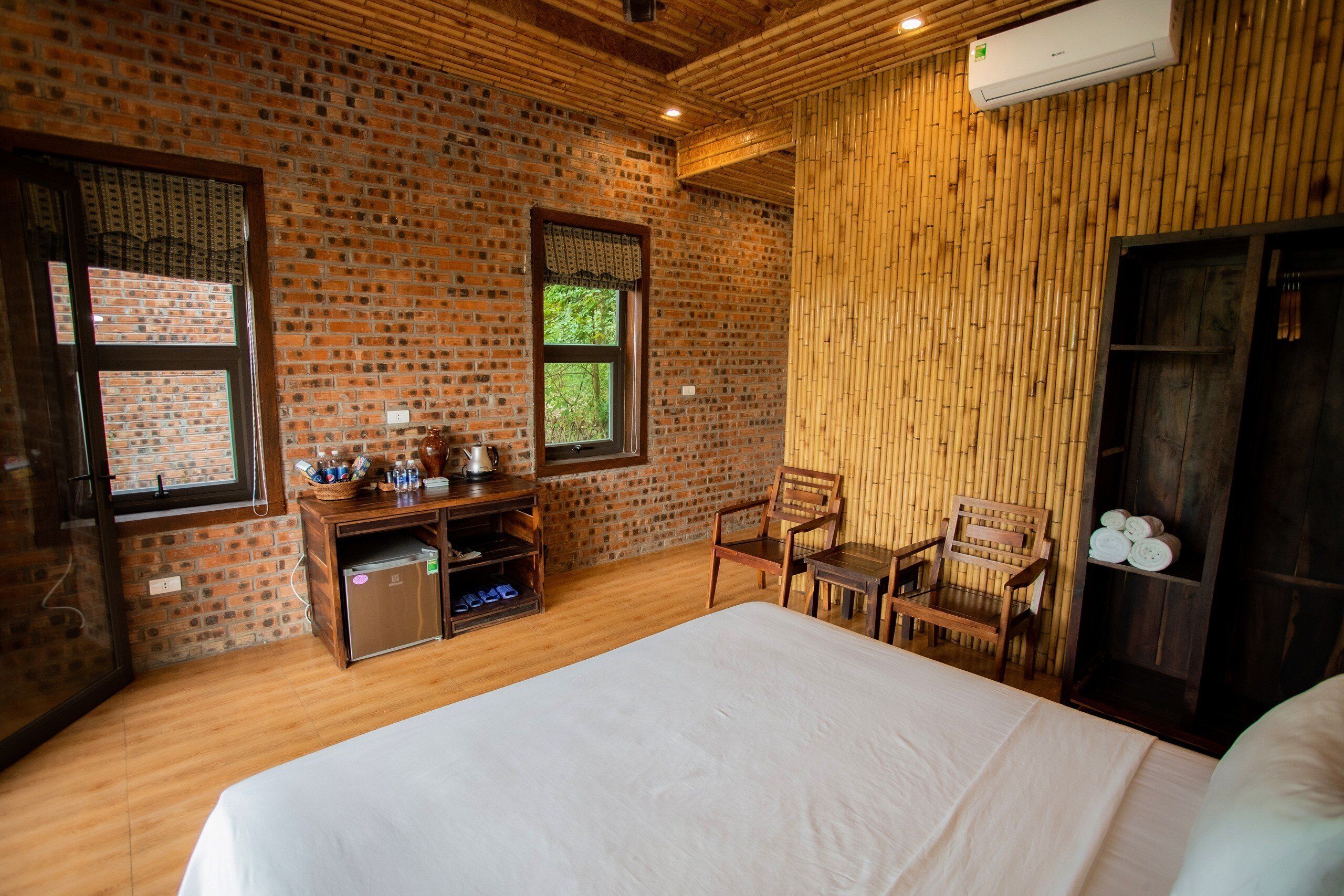 A room with a brick wall, wooden floor, a bed, a small kitchenette, and a window with a view of trees.