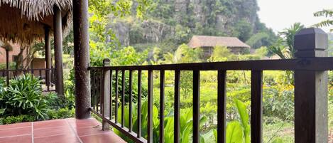A terrace with a thatched roof, red tiles, and a metal railing overlooking a lush garden and a mountain.