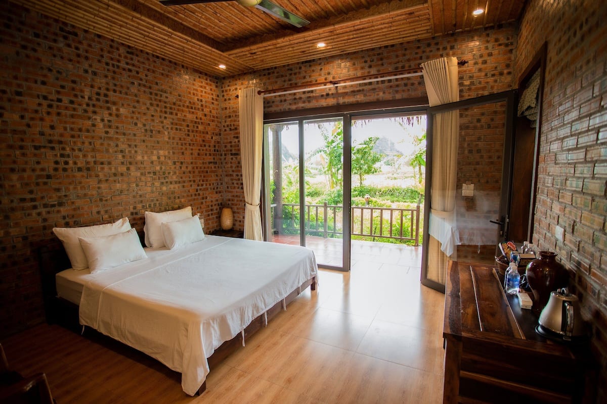 A spacious bedroom with a large bed, wooden flooring, and a brick wall. There is a balcony with a glass door leading to an outdoor area.