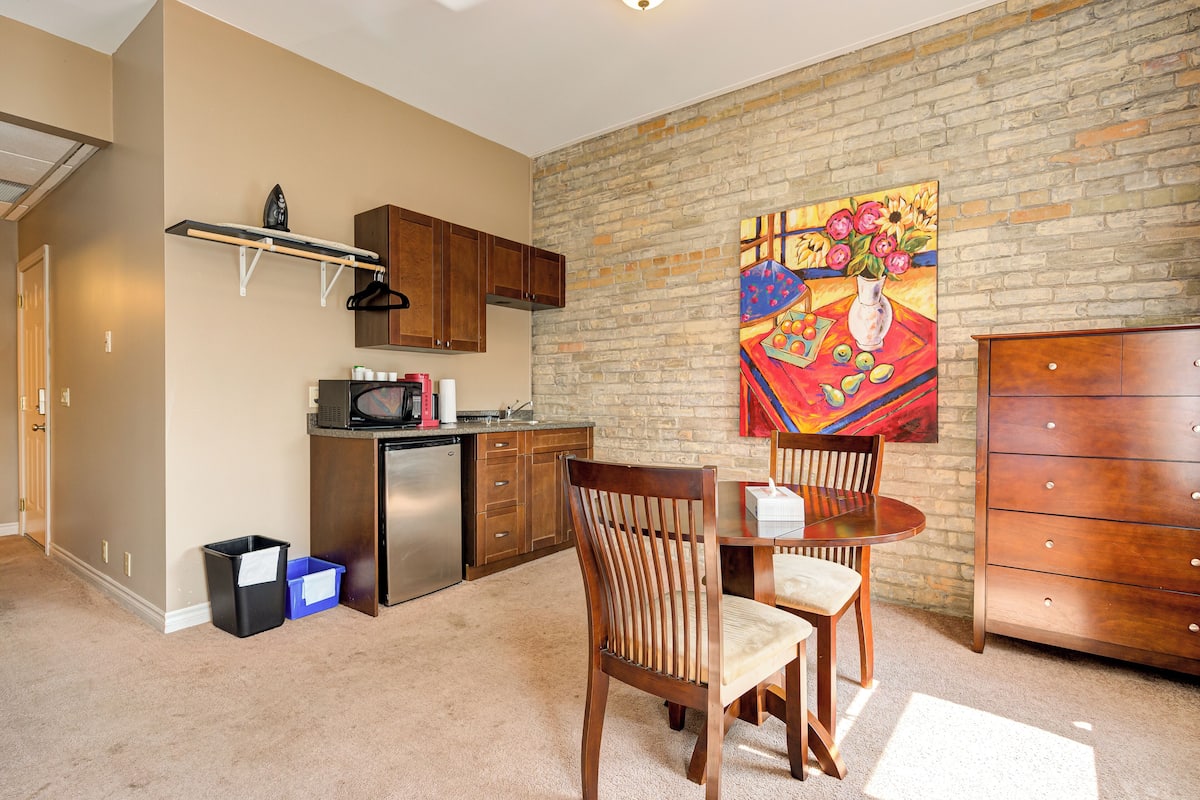 A compact kitchen with a microwave, toaster, and cabinets. There is a dining area with a table and chairs, and a painting on the wall.
