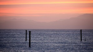 Mar tranquila con un atardecer, montañas distantes y dos pilotes en el agua.