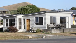 Una casa móvil de un piso con un porche delantero, un pequeño patio con muebles de exterior y una colina al fondo.