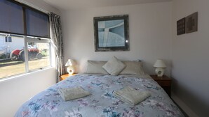 Un dormitorio con colcha estampada, dos almohadas, una mesita de noche con lámpara y una ventana con cortinas.