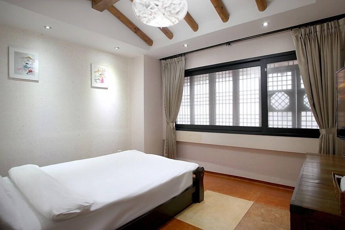 Un dormitorio con una cama individual, un escritorio de madera, un televisor y un ventanal grande con cortinas.