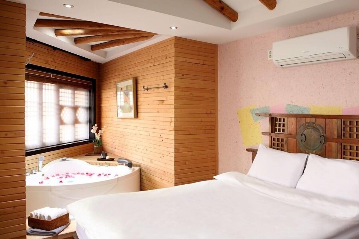 Una habitación con techo de listones de madera, una cama grande con ropa blanca, una bañera con pétalos de rosa y un aire acondicionado instalado en la pared.