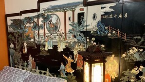Quarto tradicional chinês com um grande mural, uma cama com roupa de cama estampada e uma pequena mesa com um candeeiro.