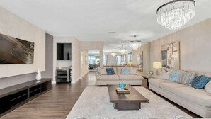 Un salon moderne avec un grand canapé, une table basse et un lustre. Un téléviseur à écran plat est fixé au mur, et il y a une cheminée.