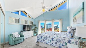 Ein Schlafzimmer mit einem großen Bett, einem Deckenventilator, einem Fernseher, einem Schminktisch und Blick auf Gebäude durch große Fenster.