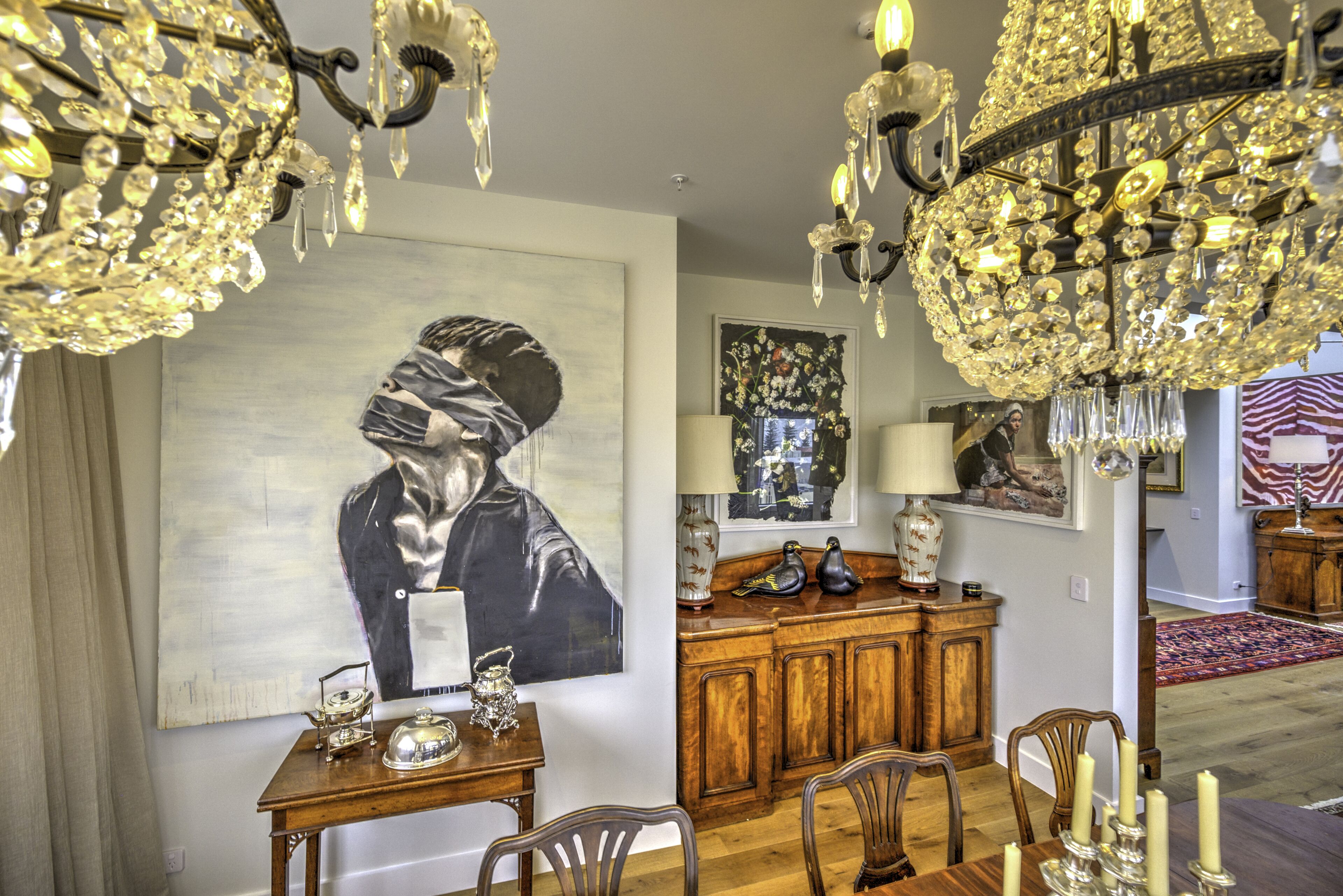 A dining area with a wooden table, chairs, and a chandelier. There is a painting on the wall and a cabinet with decorative items.