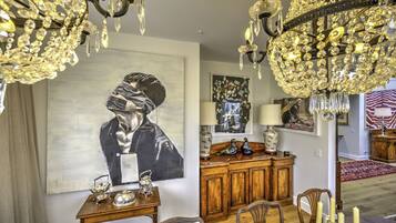 A dining area with a wooden table, chairs, and a chandelier. There is a painting on the wall and a cabinet with decorative items.