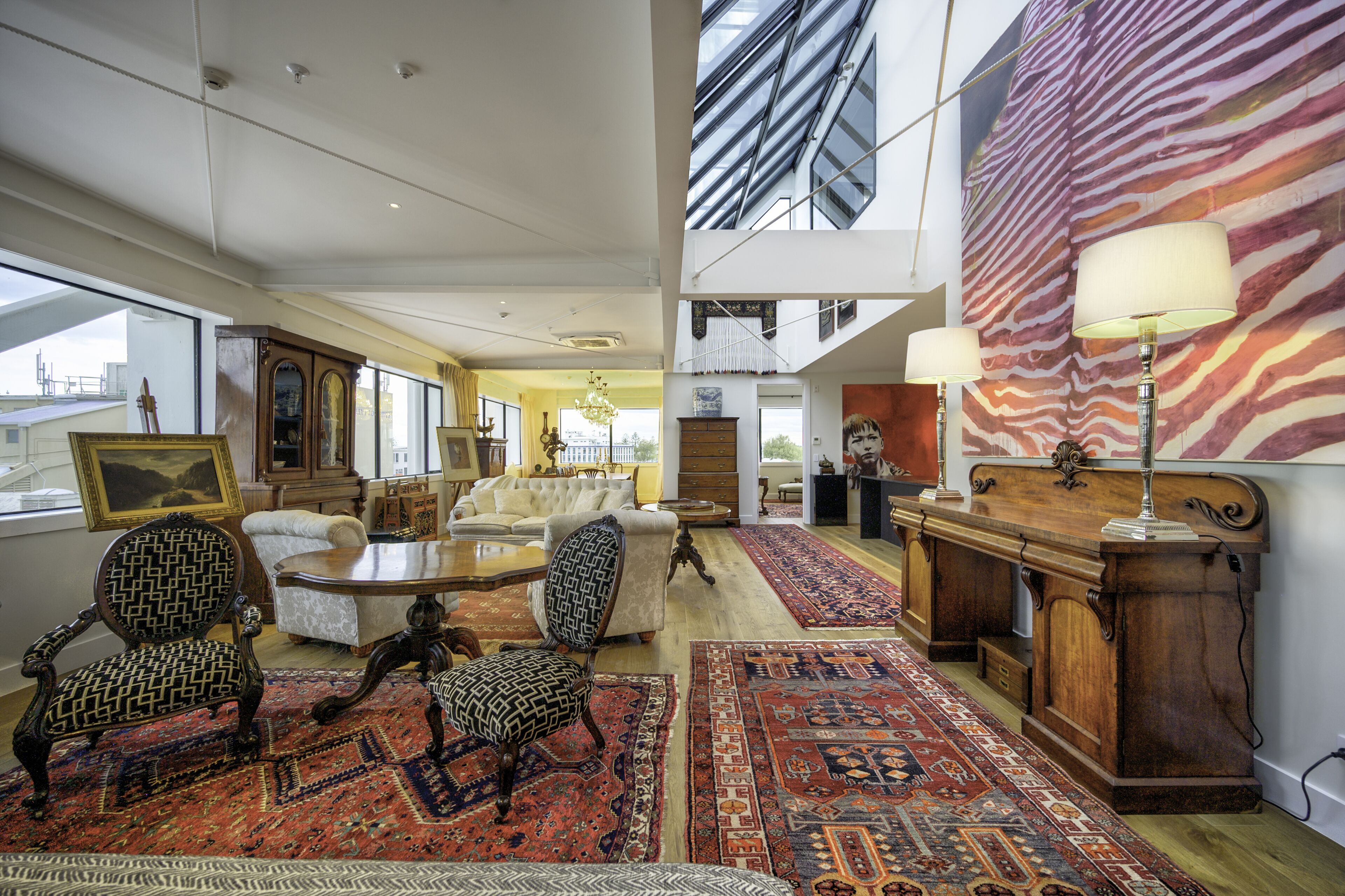 A spacious living room with a high ceiling, large windows, and a view of the sky. There are multiple seating areas, including a sofa and chairs, with a large rug on the floor.