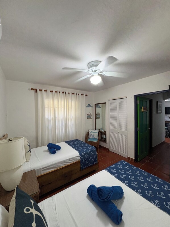 A room with two beds, a ceiling fan, a window with curtains, a chair, a mirror, and a door leading to another room. - Reefside Villa Tobago (Bon Accord)