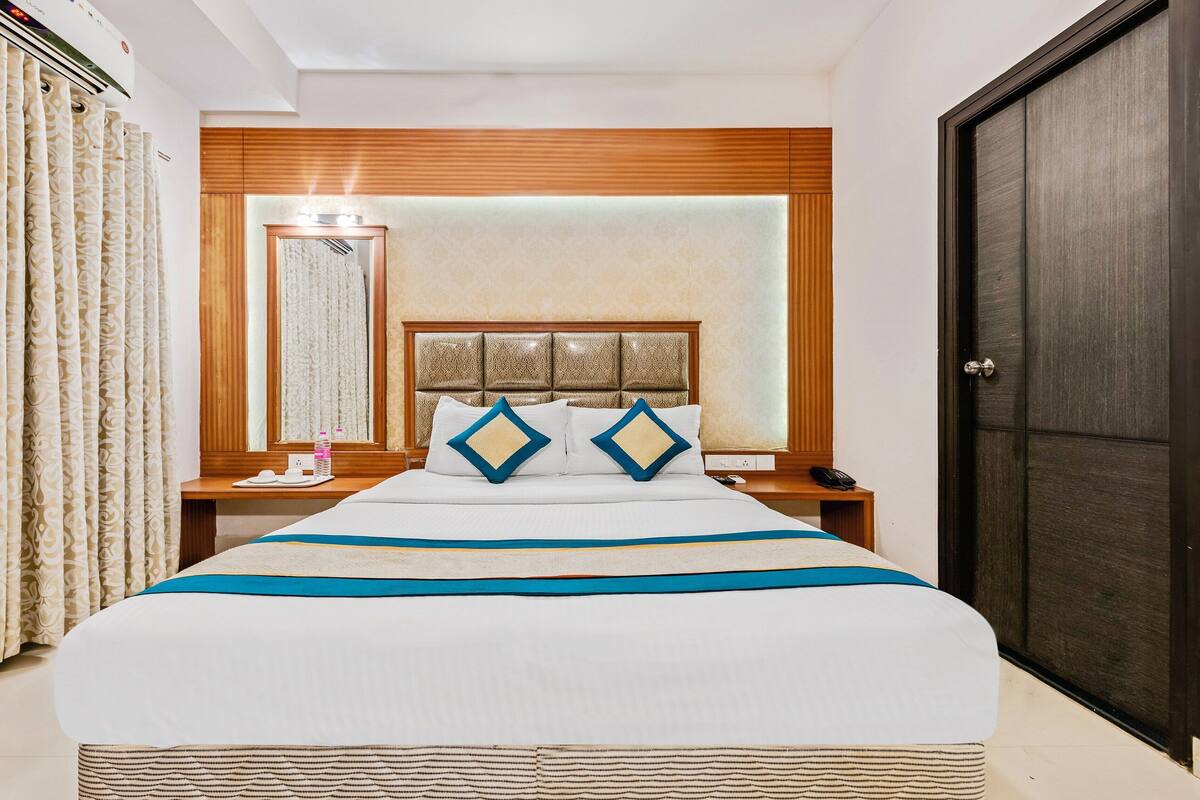 A hotel room with a large bed, a bedside table with a lamp, a nightstand with a phone, and a dark door.