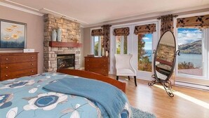 Une chambre avec une cheminée en pierre, un lit recouvert d’une couvre-lit à motifs floraux, un fauteuil et une vue sur un lac, visible à travers de grandes fenêtres.