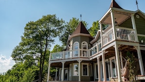 Une grande maison à deux étages, dotée d’une véranda qui fait le tour de la maison, d’une tourelle et d’un balcon couvert.