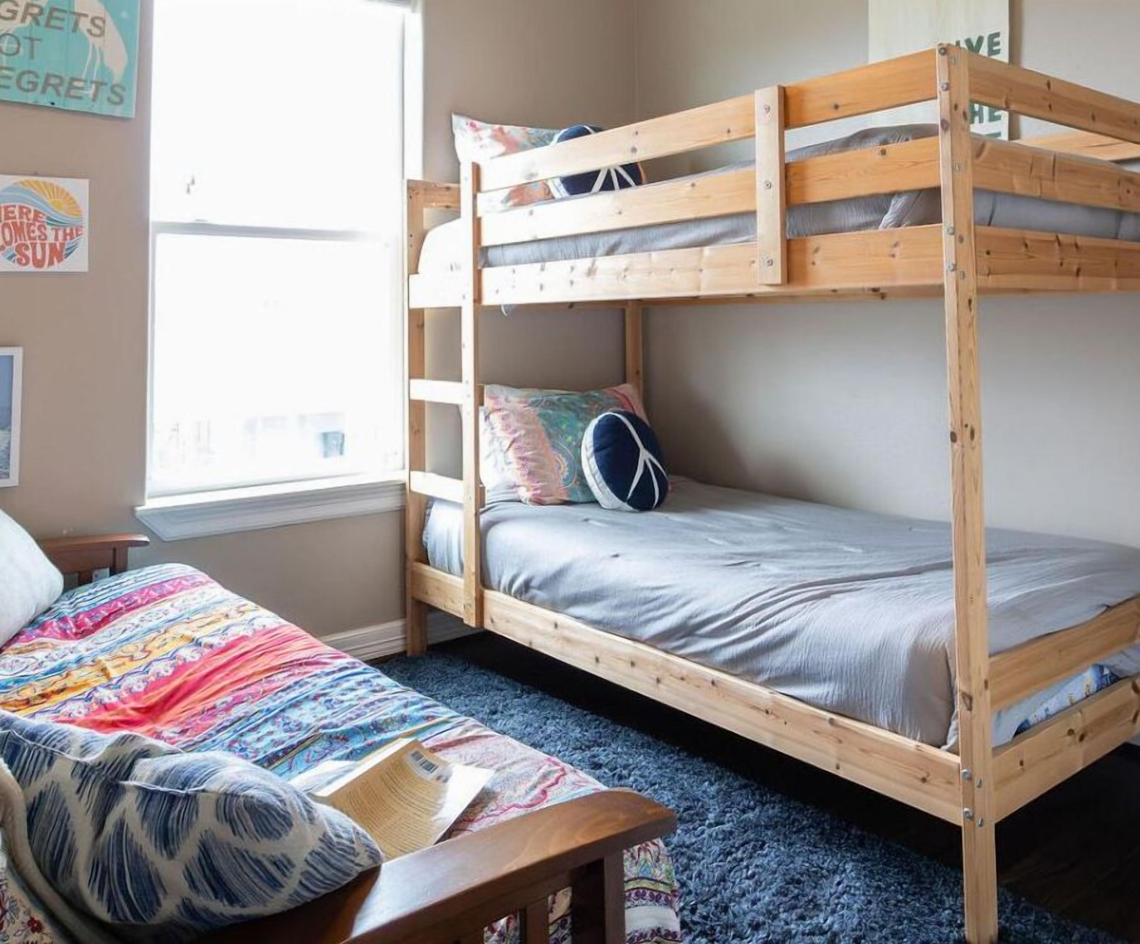 Un letto a castello con un letto inferiore e un letto singolo, entrambi con biancheria grigia. Una coperta colorata è sul letto inferiore. Una sedia di legno è di fronte al letto.