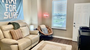 A living room with a beige couch, a patterned rug, a neon sign, and a window with blinds. - Best of Tides - Make family memories! (Galveston)