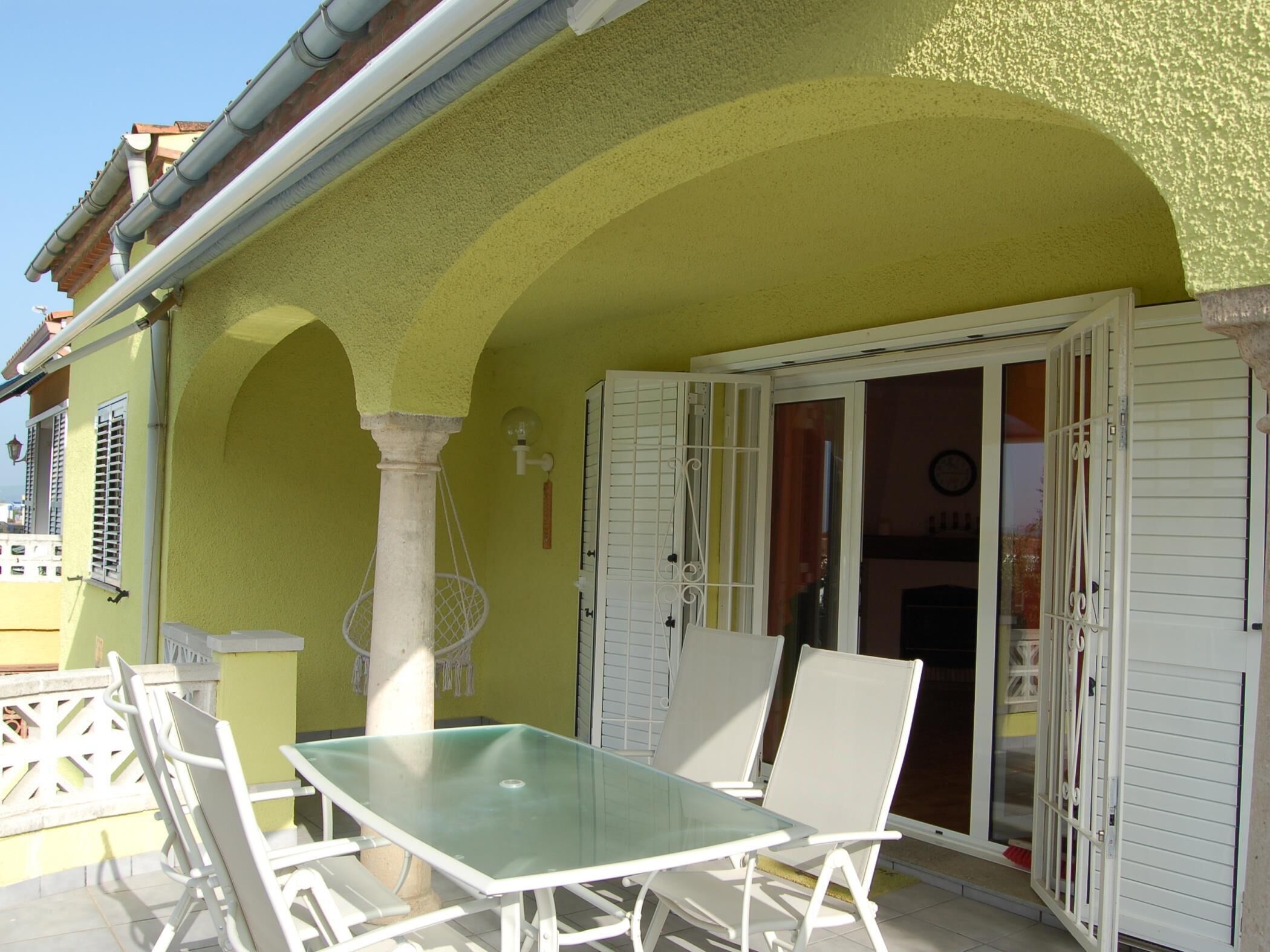 A patio with a glass table and white chairs, a yellow wall, and a door with white shutters.