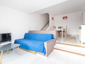 A modern living room with a blue sofa, a flat-screen TV, and a dining area with a table and chairs.