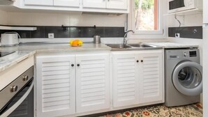 Une cuisine compacte avec des meubles blancs, un four encastré, un lave-linge et une fenêtre donnant sur un espace verdoyant.