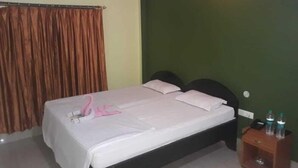 Un letto singolo con materasso bianco e due cuscini, un comodino in legno con due bottiglie, e una tenda alla finestra.