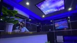 Ein moderner Empfangsbereich mit einem großen digitalen Bildschirm, der eine Naturansicht zeigt, einem Empfangstresen, einem Blumentopf und einer Sitzecke mit einem blauen Sofa.