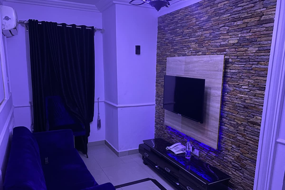 A modern living room with a dark blue sofa, a flat-screen TV mounted on a stone wall, a black entertainment unit with a phone and a bottle, and a black curtain.