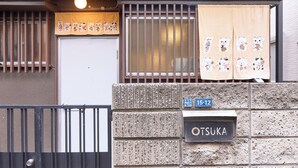 En bygning med en stenfasadde, et sort metalgitter, en hvid dør og et skilt med teksten 'OTSUKA'.