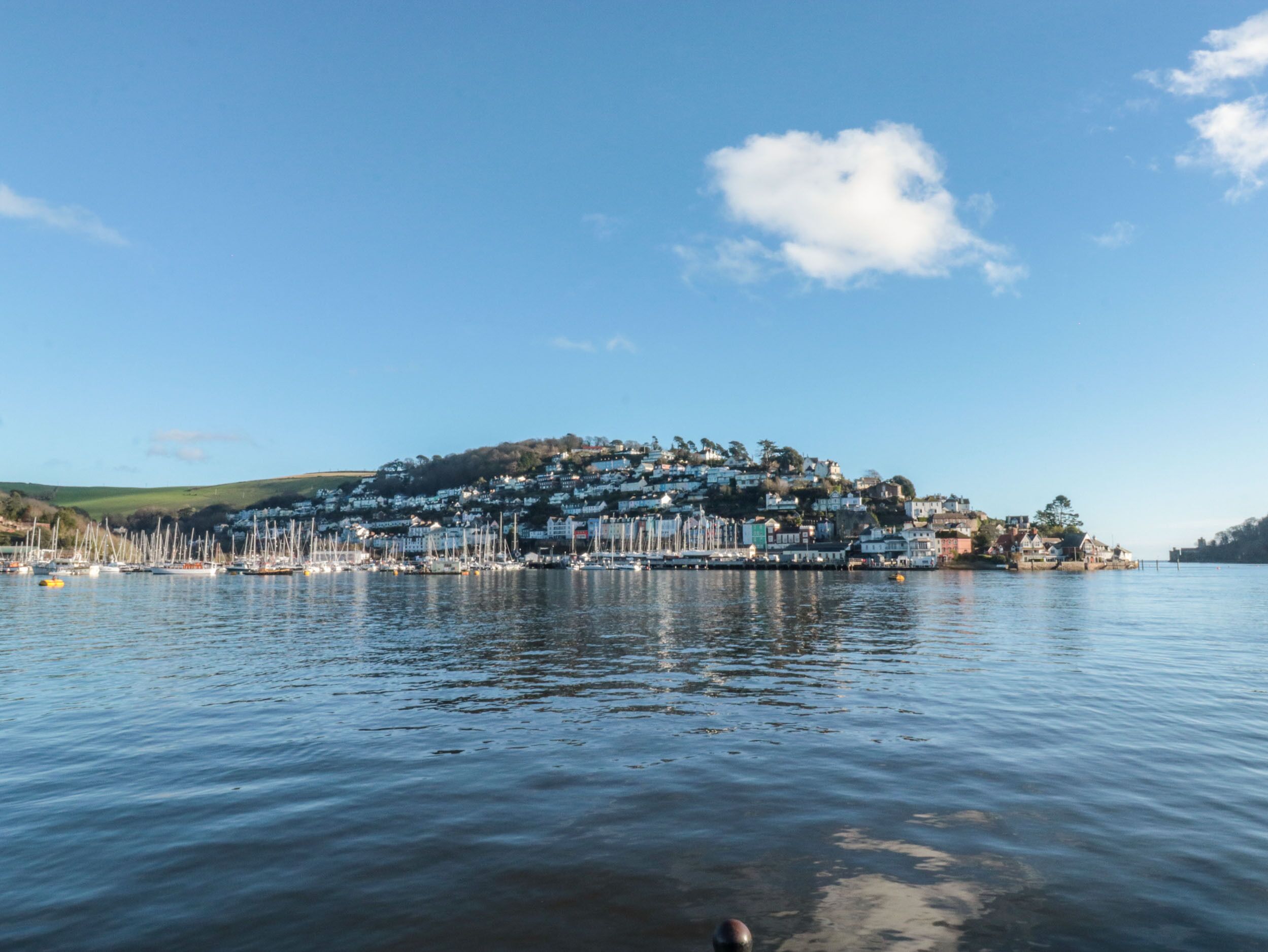 A coastal town with a marina filled with boats, a hilltop with buildings, and calm water.