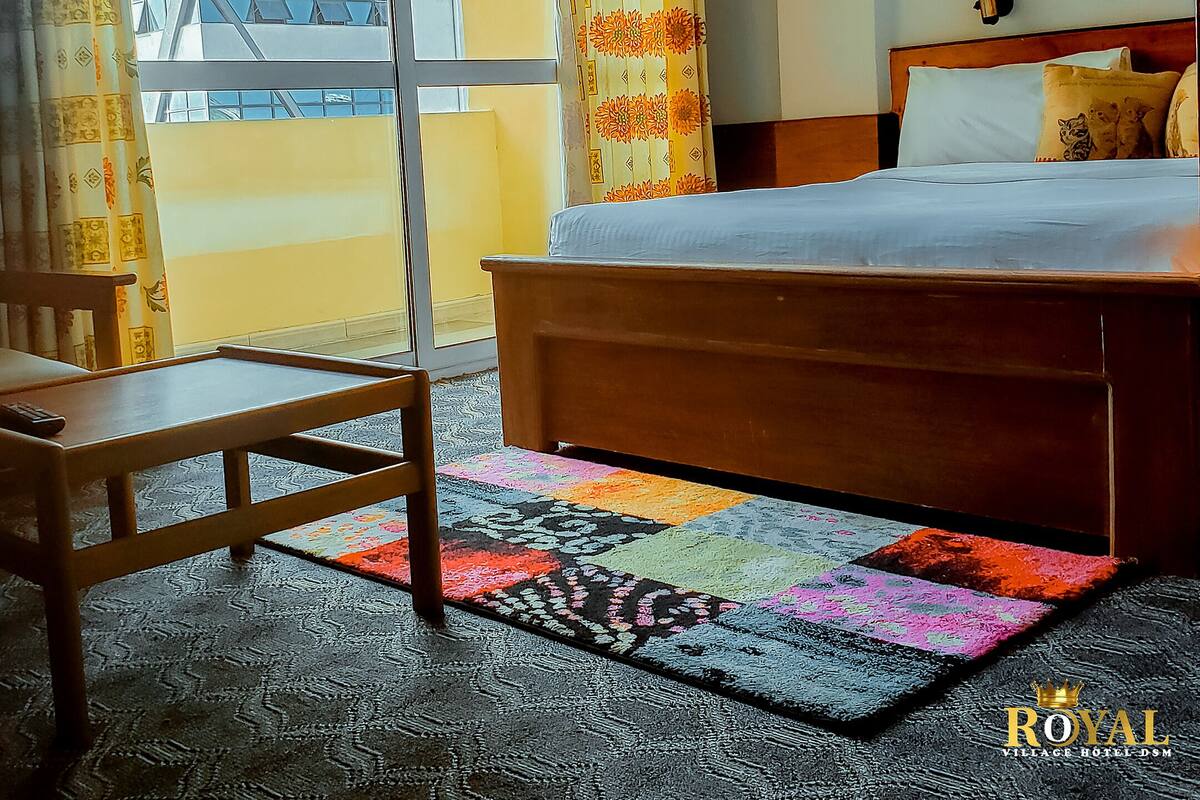 A hotel room with a large bed, a wooden nightstand, a chair, and a colorful rug.