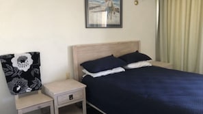 A bed with a dark blue cover, two pillows, a nightstand with a lamp, and a framed picture on the wall. - Dulkara 11 Ocean Breezes and Beachfront Cafes Nearby (Kings Beach)