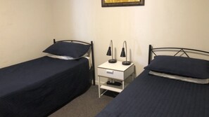 A small, simple room with two single beds, a bedside table with a lamp, and a small drawer unit. - Dulkara 11 Ocean Breezes and Beachfront Cafes Nearby (Kings Beach)