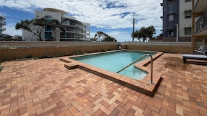 Una piscina con borde de ladrillo, rodeada por una valla, y edificios al fondo.
