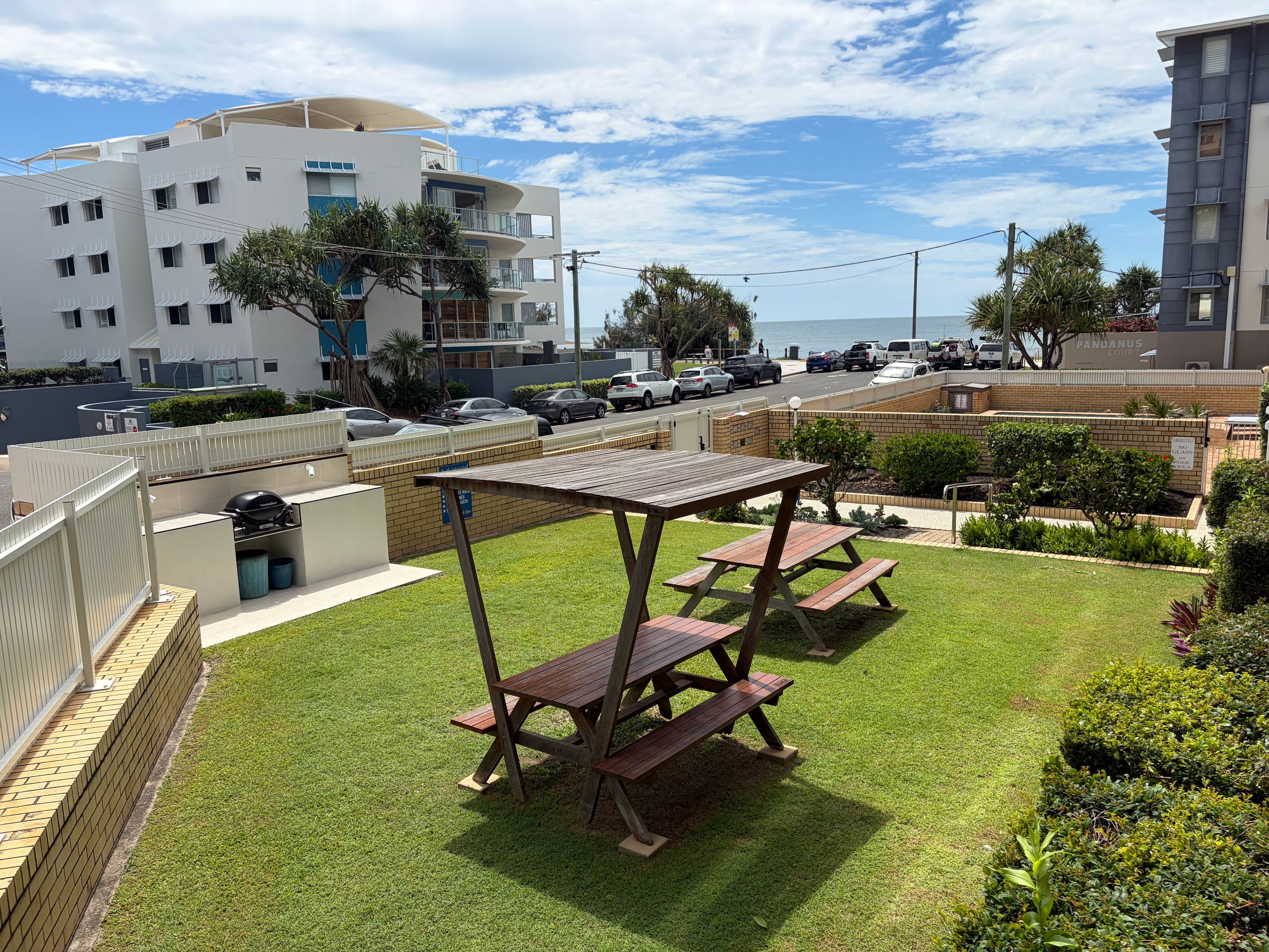 A residential area with a grassy lawn, picnic tables, a barbecue grill, and a view of the sea.