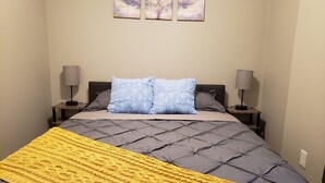 Un dormitorio con una tapicería gris capitonada en la cabecera, una manta amarilla de punto, dos almohadas grises y dos lámparas de noche.