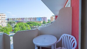 Een balkon met een kleine ronde tafel en twee witte plastic stoelen, met uitzicht op een woonwijk met kleurrijke gebouwen en groene bomen.