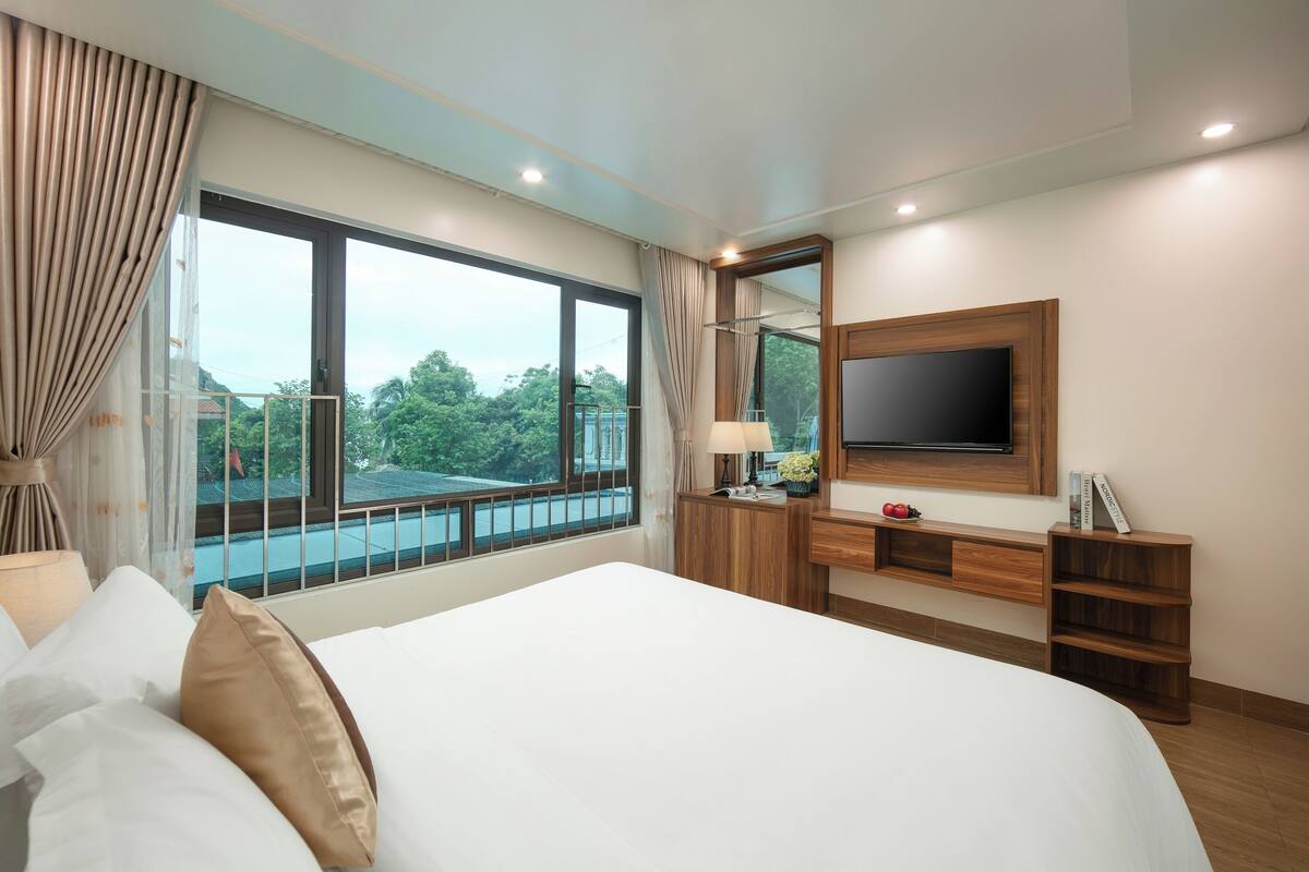 A hotel room with a large bed, a TV mounted on the wall, a wooden desk, and a window with a view of greenery.
