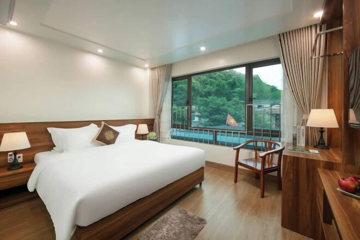 A hotel room with a large bed, a desk with a chair, a window with a view of greenery, and a balcony.