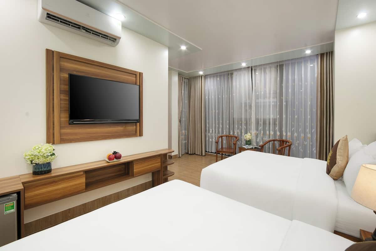 A hotel room with two beds, a TV mounted on the wall, a small table with chairs, and a window with curtains.