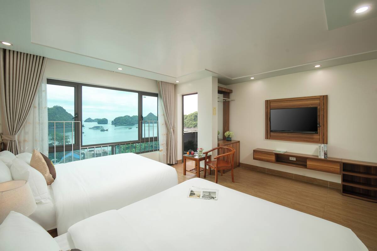 A hotel room with two beds, a TV, a desk, and a view of the sea.